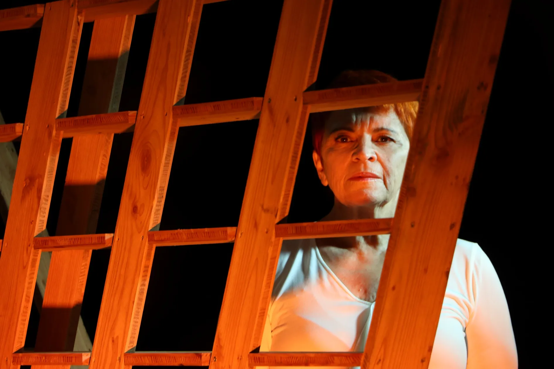 Atriz em cena observada através de estrutura de madeira, iluminação dramática a laranja, espetáculo de teatro