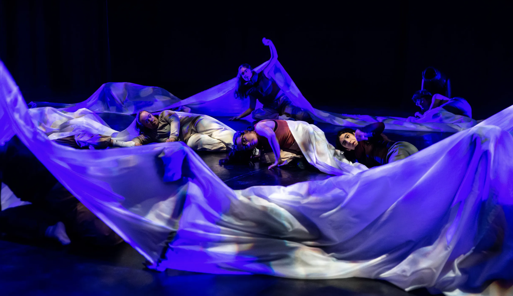 Bailarinos no chão envoltos em tecido azul flutuante, iluminação dramática