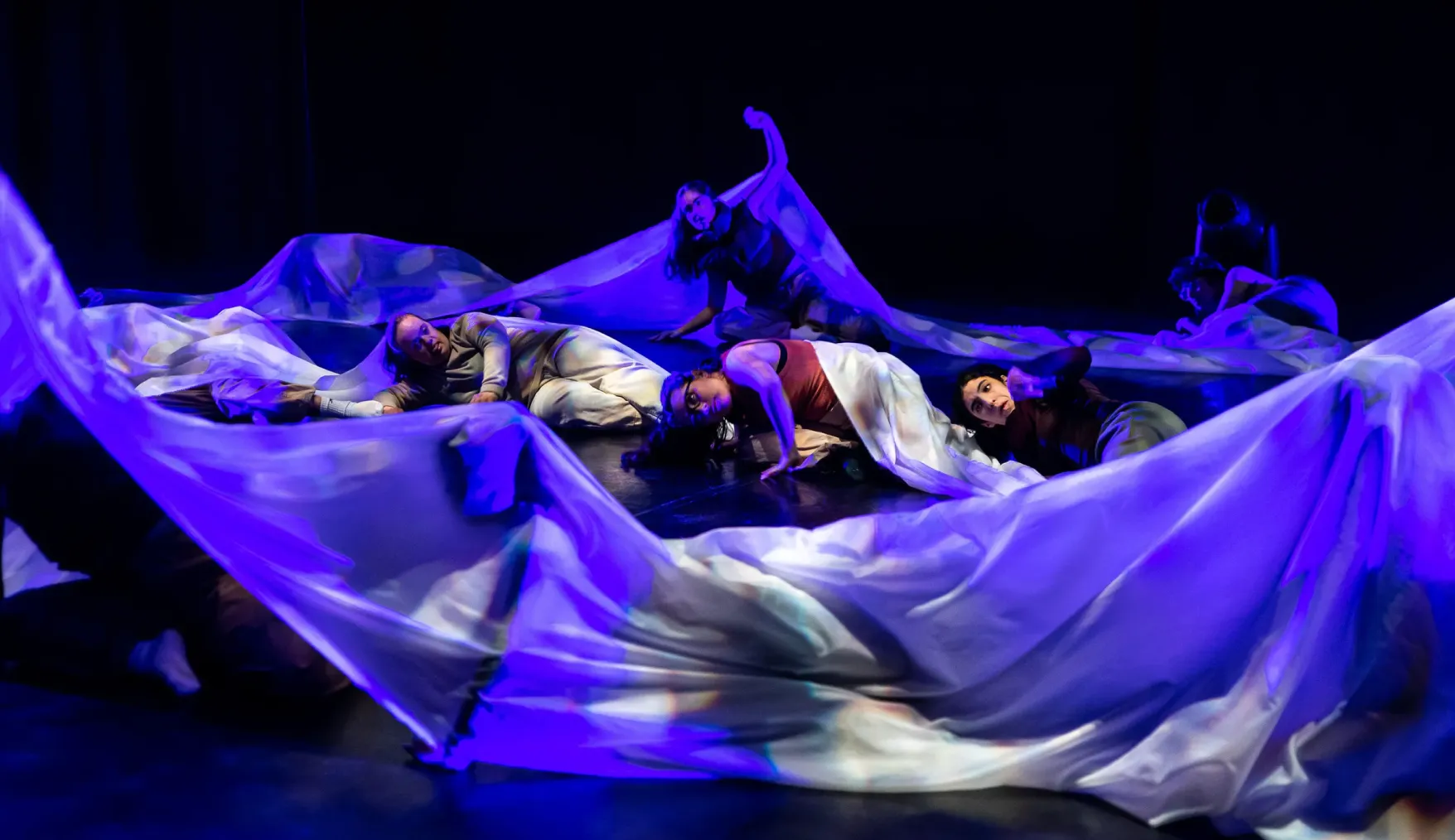 Bailarinos no chão envoltos em tecido azul flutuante, iluminação dramática