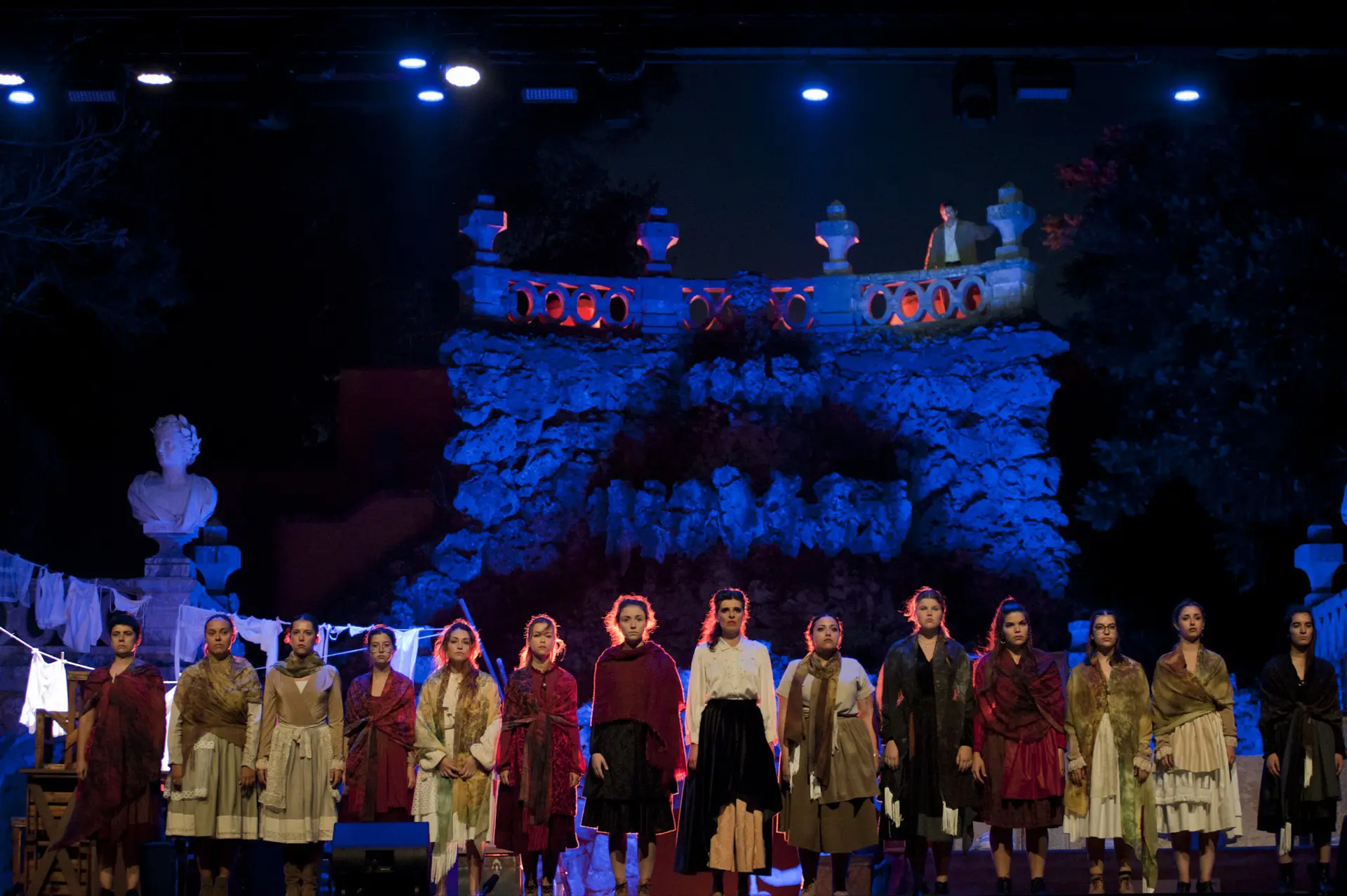 Elenco de ópera em formação num palco exterior com iluminação azul e cenário de rochedos