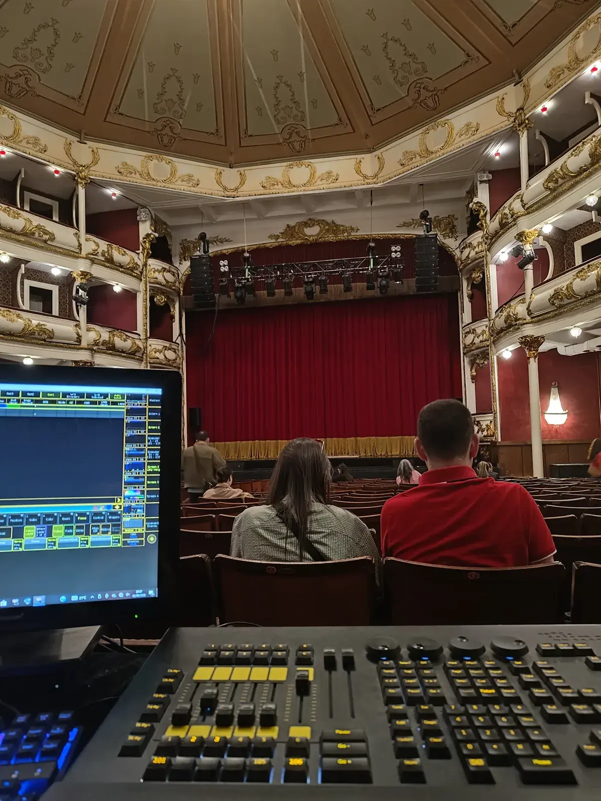 Dois técnicos na consola de luz de um teatro de ópera clássico com cortina vermelha ao fundo