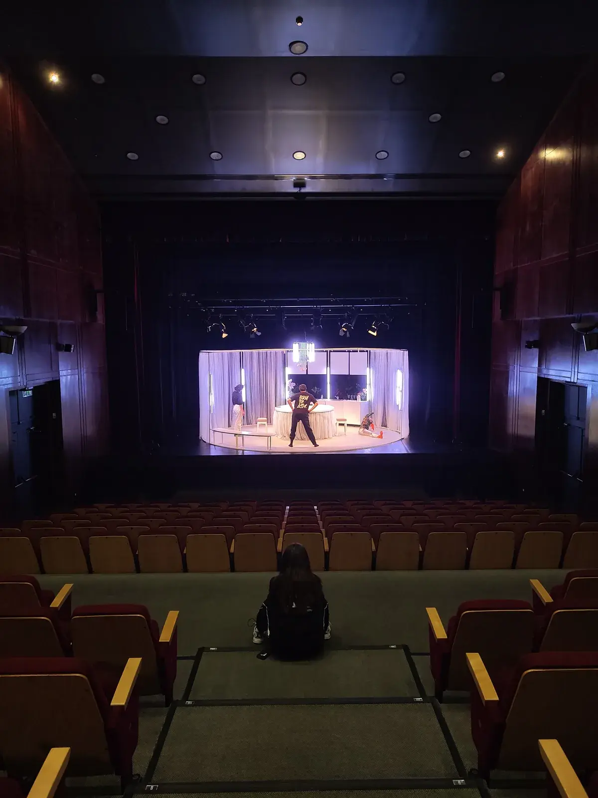 Sala de teatro vazia com palco iluminado e pessoa sozinha sentada no corredor central