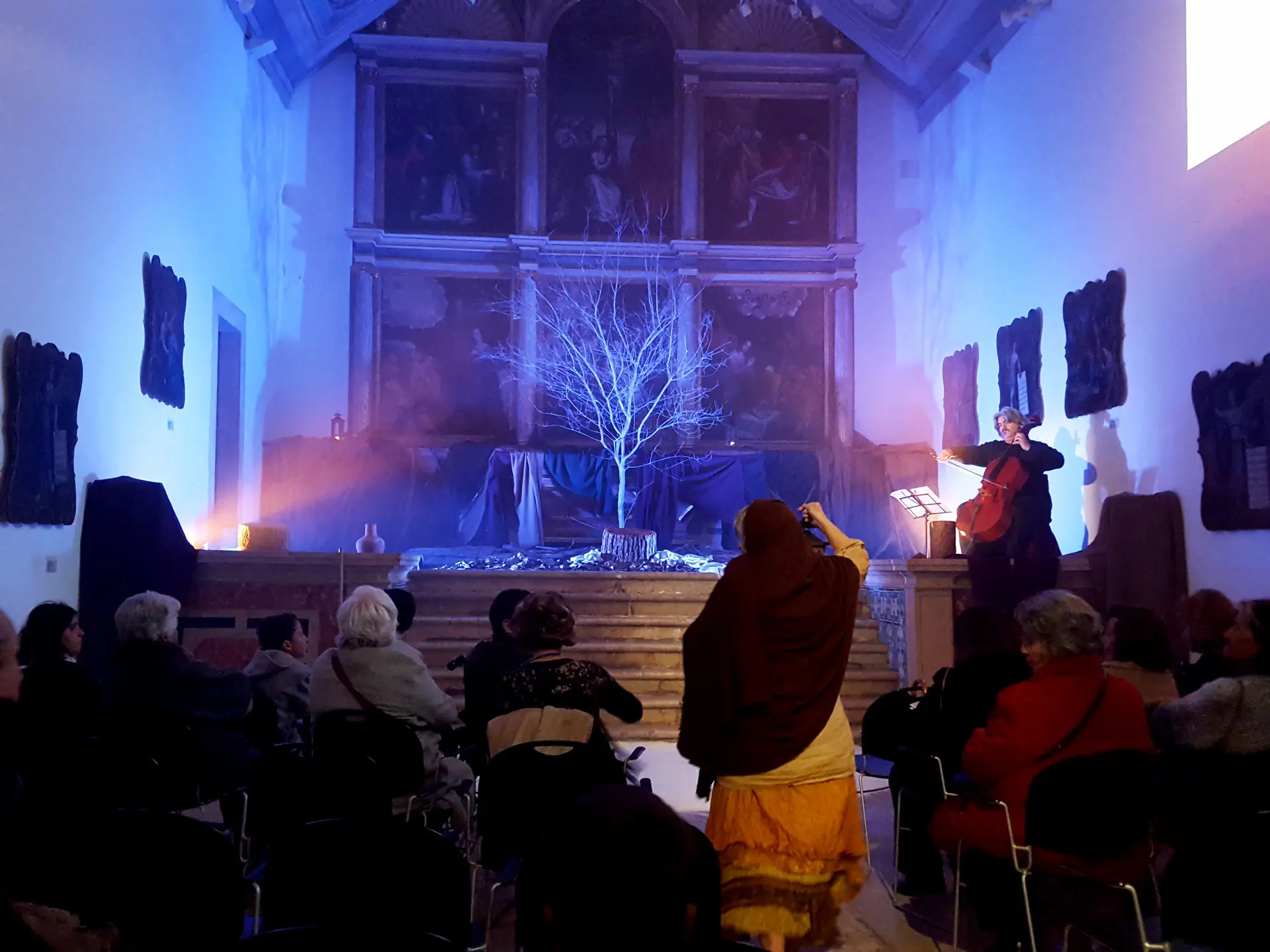 Violoncelista num altar de igreja com árvore seca iluminada a azul e público sentado